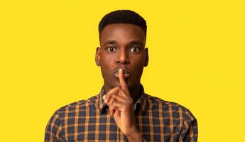It's A Secret. Young Black Guy Making Shh Silence Gesture, Mysterious African Man Putting Finger On Lips And Looking At Camera While Standing Isolated Over Yellow Background, Closeup Shot, Copy Space photo