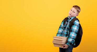 Exhausted schoolboy with backpack holding heap of books on yellow studio background, coming back home from school, copy space. Upset redhead boy have to do homework, bringing books photo