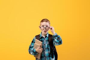 Surprised schooler with eyeglasses holding book on yellow studio background. Redhead schoolboy with backpack holding books, having amazed face expression, copy space. Books reading, education concept photo