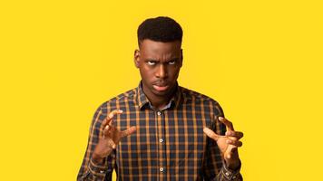 Portrait of annoyed black guy in checkered shirt emotionally gesturing, raising hands and rolling eyes with rage, feeling angry and stressed while standing isolated over yellow background, copy space photo