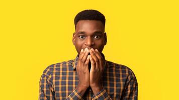Closeup portrait of black surprised guy covering his mouth with excitement and looking at camera, african american man having wonder face expression, standing isolated on yellow background, free space photo