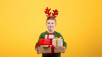 Smiling boy with festive deer accessory holding Christmas gifts over yellow studio background, empty space. Happy kid celebrating New Year, got a lot of presents, cheerfully posing at camera photo