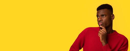 Doubtful Young Black Man Looking Aside At Copy Space With Curiousity. Unsure African Guy Touching Chin, Thinking About Interesting Offer While Standing Isolated Over Yellow Background, Panorama photo