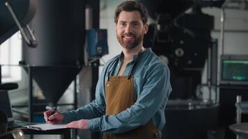 Caucasian man working in factory roasted coffee beans writing paper note check roasting machine male inspector technologist industry male guy in apron smiling looking at camera professional barista video
