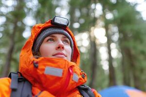 Adventurous woman wearing bright orange outdoor gear and headlamp, gazing upwards in a forest, surrounded by tall trees and a colorful camping tent, embodying the spirit of exploration photo
