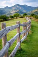 Wooden fence lines a lush green field, leading towards distant mountains under a partly cloudy sky, capturing the essence of rural tranquility and natural beauty photo