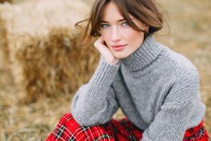 Young woman with brown hair wearing a cozy gray sweater and plaid skirt, sitting on hay bales in a rustic outdoor setting, exuding warmth and natural beauty photo