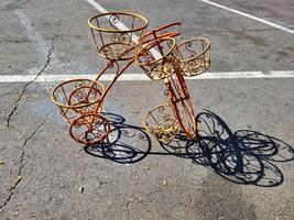 An orange empty decorative wrought-iron flower stand in the shape of a bicycle with baskets for plants, stands on the sidewalk photo
