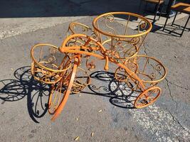 An orange empty decorative wrought-iron flower stand in the shape of a bicycle with baskets for plants, stands on the sidewalk. photo