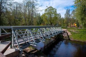 A modern metal pedestrian bridge crosses a calm waterway, connecting grassy areas. A park with slides, swings, and green trees is visible under a sunny sky. photo