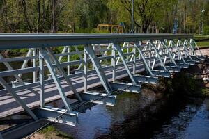 A metal lattice style pedestrian bridge spans a narrow waterway, reflecting in the water. Nearby are trees, a playground with swings, and park features. photo