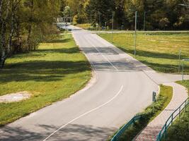 A curved road bordered by grass fields and trees, with a pedestrian pathway on the right. Streetlights line the road, and sunlight casts shadows. photo