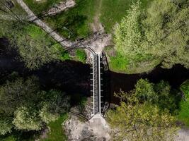 An overhead view shows a pedestrian bridge with metal railings and stone supports crossing a narrow river, surrounded by green trees and pathways. photo
