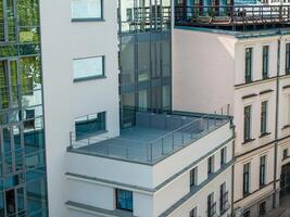A modern building with large glass windows and a terrace stands beside a traditional cream colored structure with ornate detailing. photo