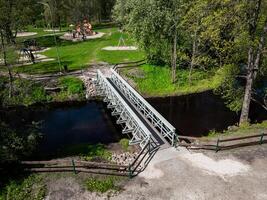 A small metal bridge with wooden railings crosses a narrow stream in a park. The park includes a playground, grassy areas, trees, and visible pathways. photo