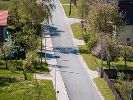 Quiet residential street lined with trees, small houses with red and dark roofs, patterned sidewalks, streetlights, power lines, and blooming gardens. photo