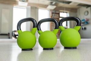 Three vibrant green kettlebells with black handles are aligned on a polished surface in a gym. Blurred shelves, a clock, and equipment are visible. photo