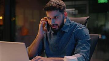Indian man businessman business entrepreneur smiling guy smiling Arabian businessman male employer in dark night evening office talking mobile phone call pleasant conversation chatting talk consulting video