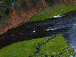 A person in a blue jacket kayaks on a dark river bordered by green grass and a red sandstone cliff with sparse trees and exposed roots. photo