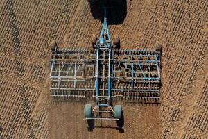 Aerial perspective of blue farming machinery attached to a tractor, tilling a field with evenly furrowed soil and parallel lines visible. photo