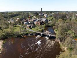 Elevated perspective of an industrial area with old brick buildings, a tall smokestack, a river dam with whitewater, and scattered trees and structures. photo