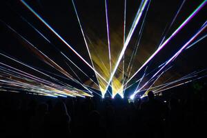 Multicolored laser beams radiate dynamically in the night sky, with silhouettes of people in the foreground and faint tree outlines on the sides. photo