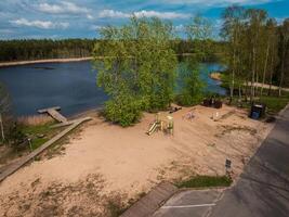 A sandy playground with a slide and swings sits near a calm body of water, bordered by wooden walkways, reeds, and trees with fresh green leaves. photo