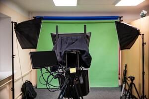 Studio setup featuring a green screen, softbox lights, teleprompter on a tripod, cables, and additional equipment in a beige carpeted room. photo