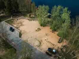 A sandy area near water with a playground, wooden dock, trees, pathways, benches, a paved parking lot, and a mix of open and wooded spaces. photo