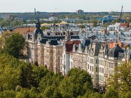 Row of ornate historic buildings with steep roofs, trees in the foreground, and modern buildings in Riga, Latvia photo