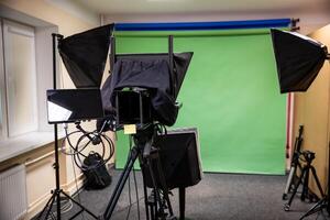 A professional studio setup with a green screen, camera on tripod, softbox lights, monitor, cables, beige walls, window, radiator, and carpeted floor. photo