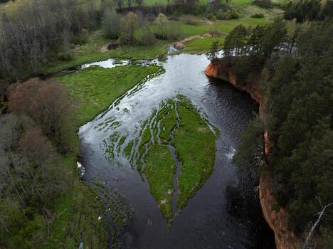 A river flows around a triangular grassy island, bordered by a red sandstone cliff with trees. Open fields and forested areas surround the scene. photo