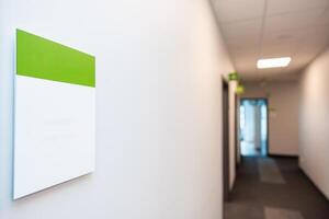 Well lit hallway with white walls, green and white sign, carpeted floor, multiple doorways, and a glass door at the end allowing natural light. photo