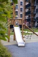 Modern playground featuring a metal slide, wooden frame, and climbing net, surrounded by pathways, greenery, and multi story apartment buildings. photo