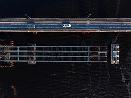 An aerial view shows a drawbridge with one section raised, vehicles on the roadway, a person in red walking, and dark water beneath the structure. photo