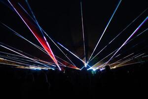 Vibrant laser beams form dynamic patterns in the dark night, with silhouetted people in the foreground and faint structures or trees in the distance. photo
