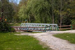 A small metal pedestrian bridge with lattice design spans a narrow waterway, surrounded by trees, grass, and yellow dandelions in a park setting. photo