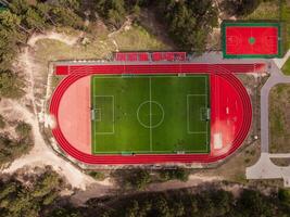 Aerial view of a sports complex featuring a red running track, green soccer field with goalposts, adjacent court, trees, pathways, and seating area. photo