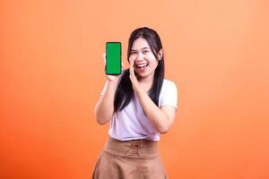 A woman asia smiling while holding a smart phone cellphone with a green screen. Hands next to her mouth announcing something wearing a white t-shirt and brown skirt. Isolated on an orange background photo