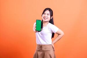 Smiling woman holding out mobile phone with green screen in front of orange backdrop photo