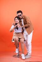 a pair of young lovers cheerful expression where the man is standing and the woman is sitting on a white chair, hands forming a sign of love very romantic. isolated on orange background photo