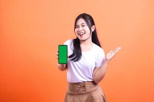 A woman holds a smart phone gadget with a green screen isolated on an orange background. She smiles and gestures as if she is presenting or demonstrating something on the phone. photo