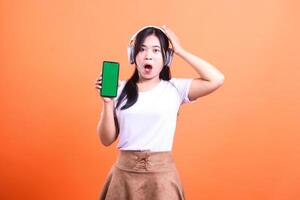 Woman holding a green screen smart phone gadget with headphone with shocked expression in an orange background photo