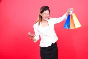 A woman in a white suit and a dark skirt holds three colorful shopping bags, she is smiling with open mouth and her both hands are at different high, isolated on a red background photo