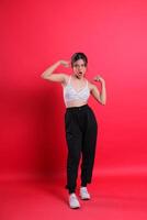 A woman wearing a white bra, black pants and white shoes shows her muscles in a flex pose, with a vibrant red studio background, isolated on a red background photo