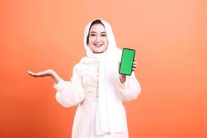 Smiling muslim woman in hijab holding up smartphone with green screen and presenting with hand gesture for copyspace. isolated on a orange background. photo
