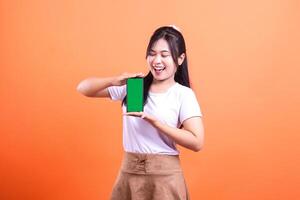 A woman is smiling while holding a smart phone device with a green screen in front of an orange background. She is wearing a white t-shirt and brown skirt. isolated on orange background photo