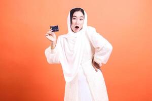 Surprised Muslim woman holding credit card with shocked facial expression, showcasing modern banking solutions and online shopping, isolated on a orange background. photo