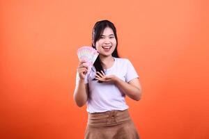 A smiling woman with a fan of currency bills, standing on an orange backdrop photo
