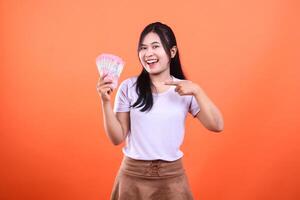 A young woman points to a pile of money, standing in front of an orange backdrop photo